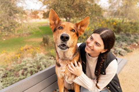 A woman and her dog sitting on a park bench. The dog is smiling at the camera