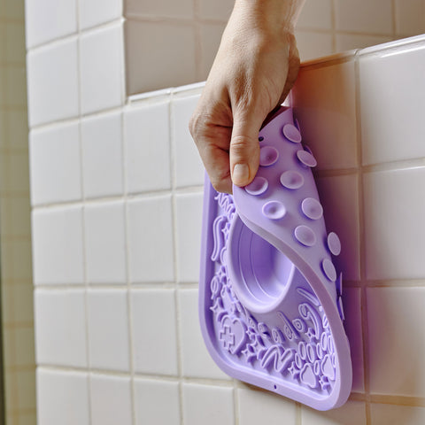 Purple silicone mat with textured design being held against a tiled bathroom wall.